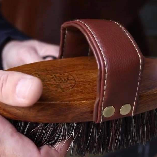 A close up view of the Hairy Pony Dandy Brush being help in a person's hand. It shows a close up view of the grain in the beechwood material as well as the soft, padded handle and bristles on the base of the brush.